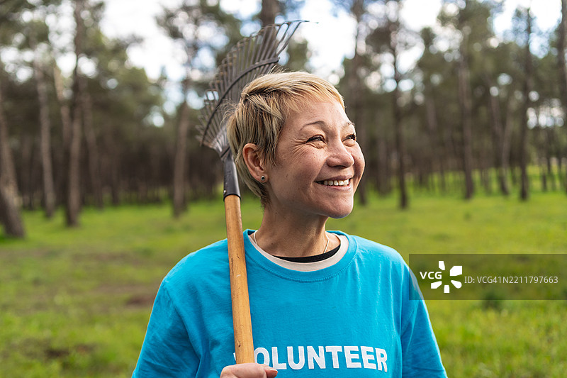 老年志愿者在社区公园里耙树叶，微笑着工作，穿着蓝色衬衫。图片素材