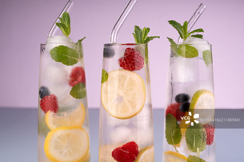 清新的柠檬、浆果和薄荷饮品三重奏，盛在高脚玻璃杯中，配有冰块和玻璃吸管，背景为柔和的紫色，营造出充满活力和清新的氛围，凉爽，具有美感。图片素材