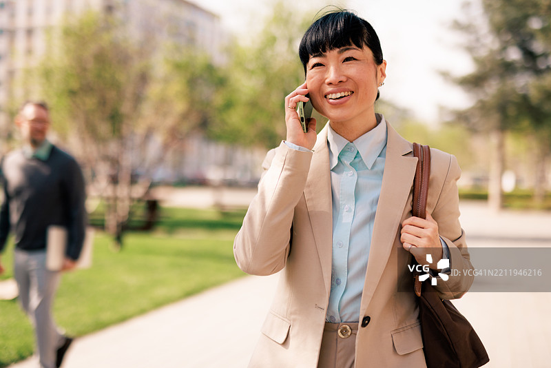 微笑的女商人在公园里走路，同时打电话。图片素材