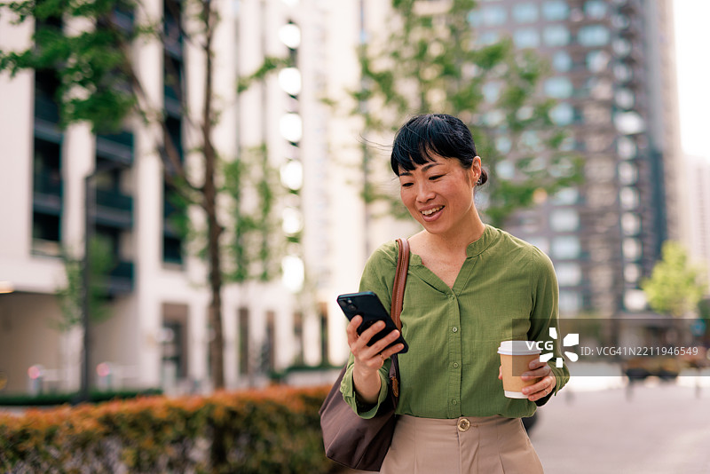 微笑的女商人在城市中走动，一边使用智能手机一边喝咖啡图片素材