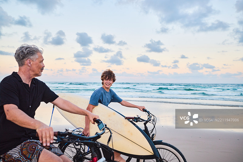 父亲和儿子在澳大利亚海滩日落时骑着电动自行车，车上放着冲浪板。图片素材