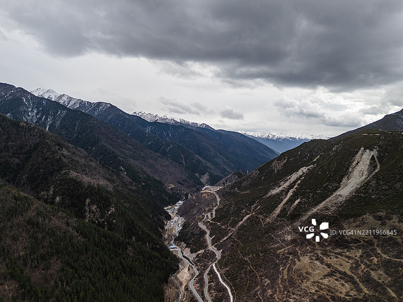 遥远村庄的鸟瞰图，坐落在狭窄的山谷中，四周被森林覆盖的山坡和白雪皑皑的山峰环绕，天空阴云密布。图片素材