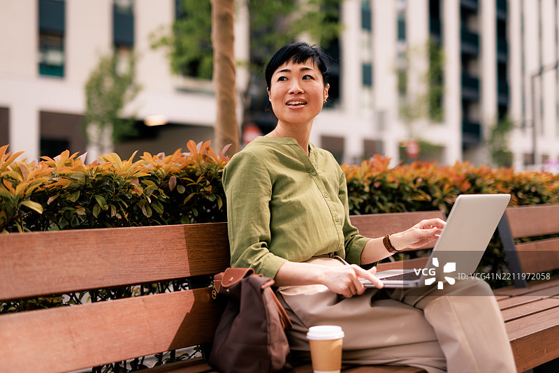 在城市的长椅上，一位女商人一边喝咖啡一边使用笔记本电脑。图片素材