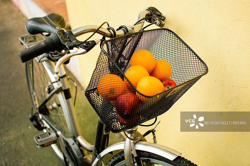 靠在黄色墙壁上的自行车，车篮里装满了新鲜的橙子和苹果图片素材