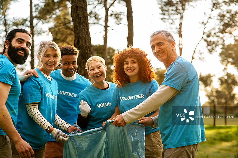 一群穿着蓝色T恤的志愿者在公园里快乐地捡垃圾，面带微笑地对着镜头，宣传社区服务。图片素材