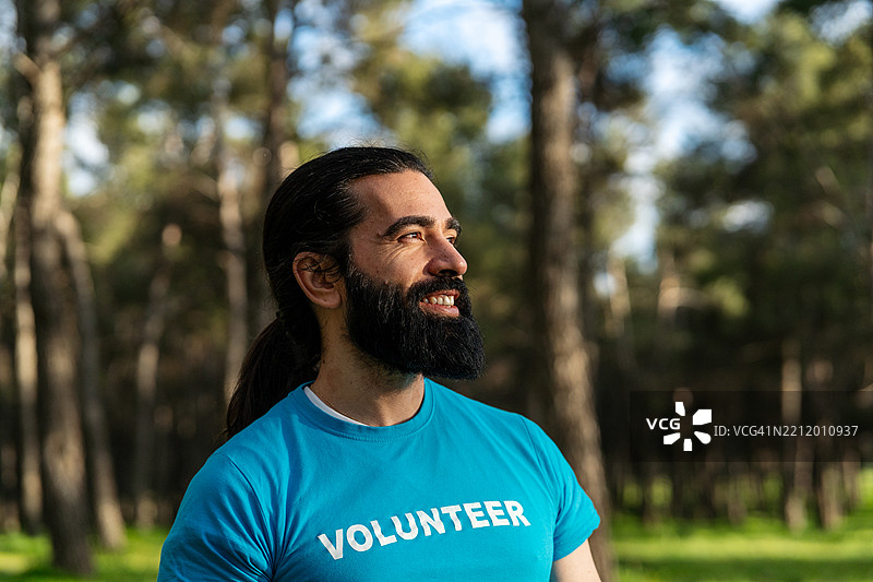 阳光明媚的森林中，一位快乐的志愿者穿着蓝色衬衫，侧身微笑。图片素材