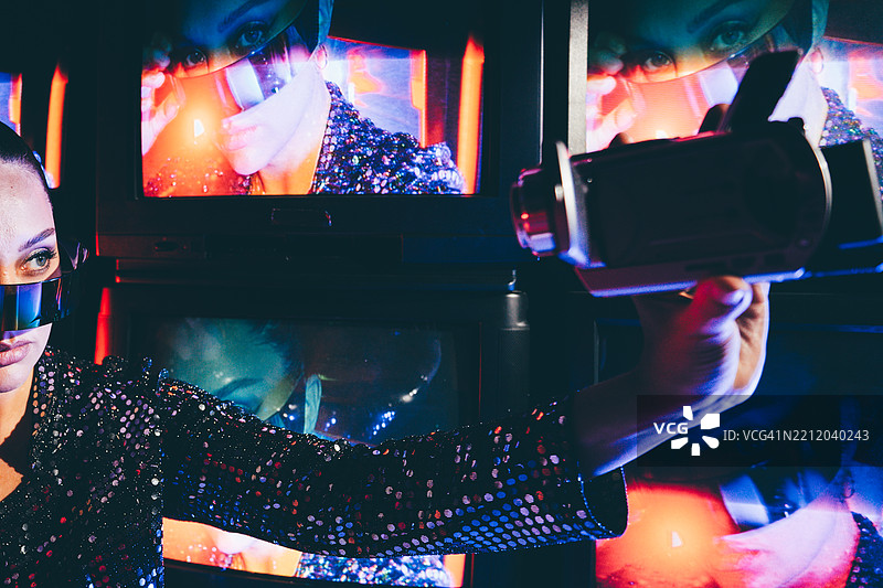 一位身着未来感亮片连衣裙、戴着紫色眼镜的女子，手持老式摄像机，在黑暗房间的多屏背景前拍摄她不同的眼睛和脸部。图片素材