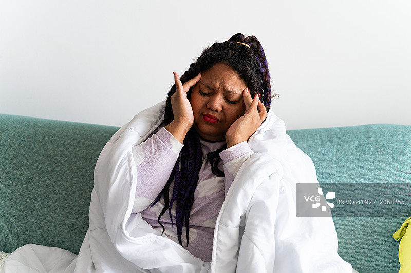 年轻女性在沙发上裹着毯子，痛苦地捂着头。图片素材