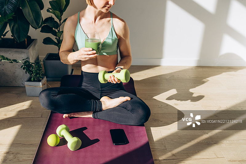 穿着健身服的女性坐在瑜伽垫上，享用绿色果昔，刚完成家庭锻炼。图片素材