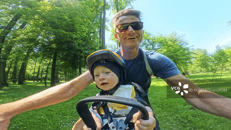 微笑的白人父亲和婴儿男孩在阳光明媚的绿色森林中一起骑自行车图片素材