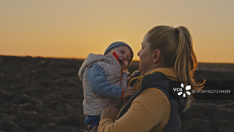 母亲在美丽的日落时分抱着她的儿子，站在刚耕耘过的田野上图片素材