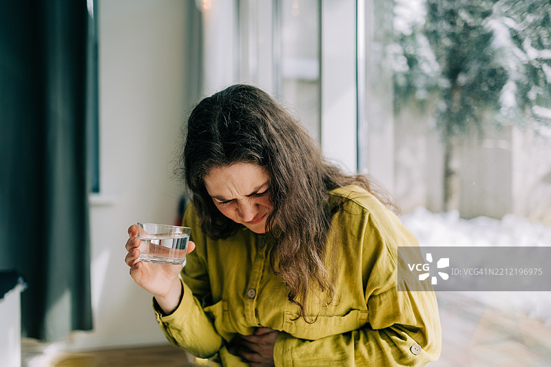 一位因腹痛而苦恼的女性在窗边拿着一杯水图片素材