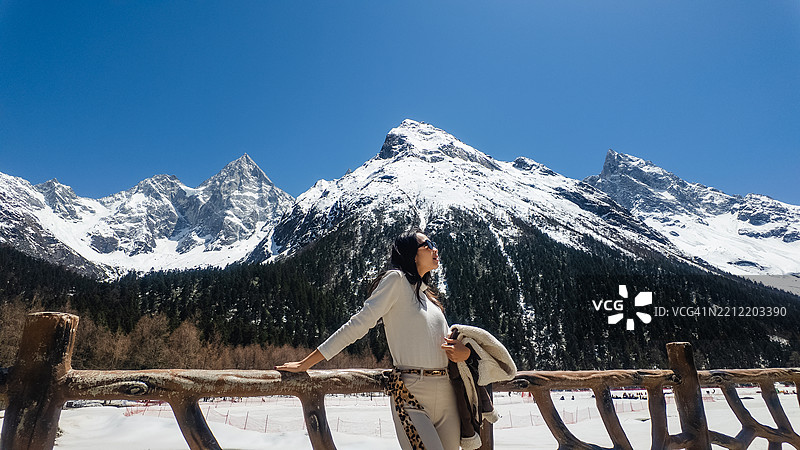 一位时尚女性穿着冬季服装，站在一座乡村风格的木栅栏旁，身后是雄伟的雪山和湛蓝的天空。图片素材