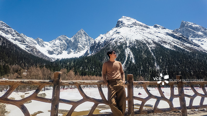 一名穿着休闲的男子靠在木栅栏上，欣赏着在蔚蓝天空下的雪山壮丽景色。图片素材