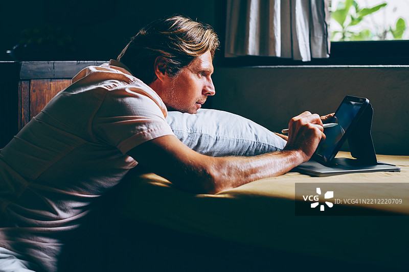 中年欧洲金发男子坐在家中的沙发旁，专注地使用带触控笔的平板电脑，窗外自然光线洒入。图片素材