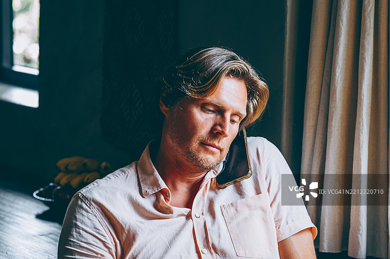 男子坐在窗边打电话，表情严肃地向下看，室内光线昏暗自然。图片素材