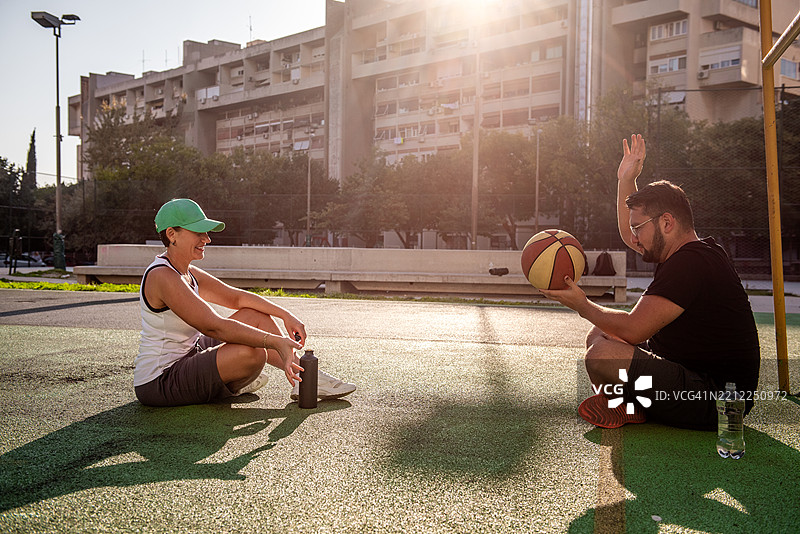 两个朋友在打完篮球后休息，坐在球场上笑着。图片素材
