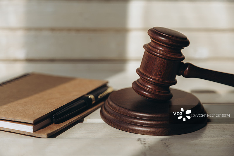 在浅色木质背景下，螺旋记事本和一支笔放在棕色法槌旁边，法槌放在声音块上。图片素材