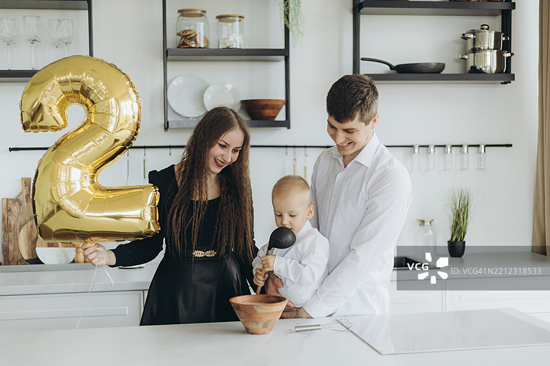 快乐的年轻女性在厨房里与丈夫和儿子一起庆祝生日，手里拿着一个金色的数字2气球。图片素材