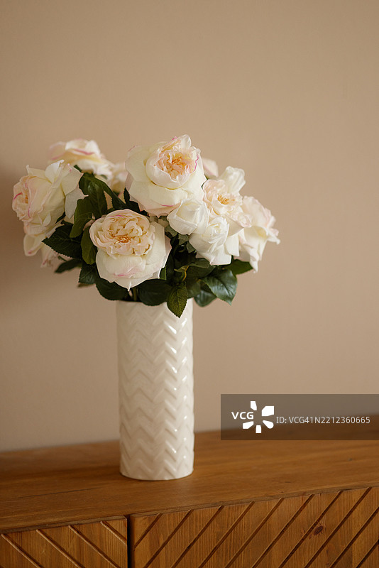 白色花瓶中盛放着柔软的白玫瑰，摆放在木质家具上图片素材