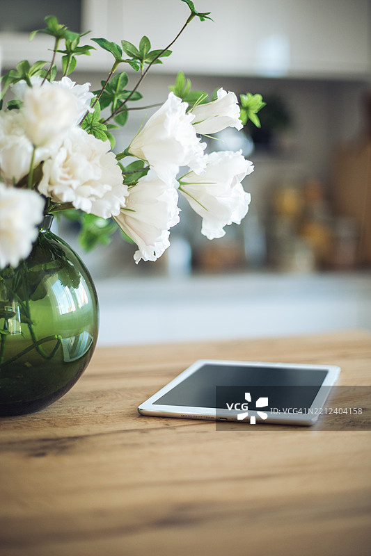 厨房木桌上，绿色花瓶里的白色花朵旁边放着一台平板电脑。图片素材