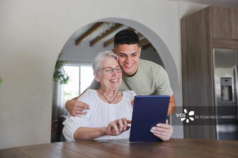 一位拉丁裔的孙子和祖母穿着休闲服装坐在厨房桌旁使用数字平板电脑。图片素材