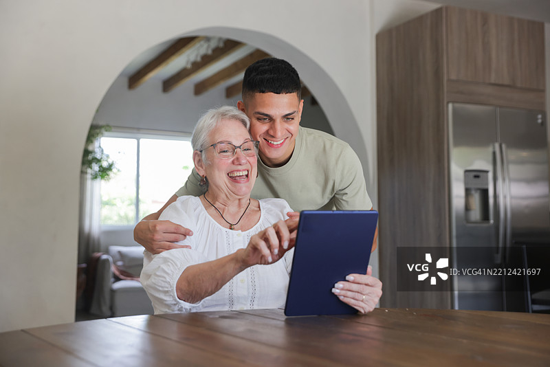 一位拉丁裔的孙子和祖母穿着休闲服装坐在厨房桌旁使用平板电脑。图片素材