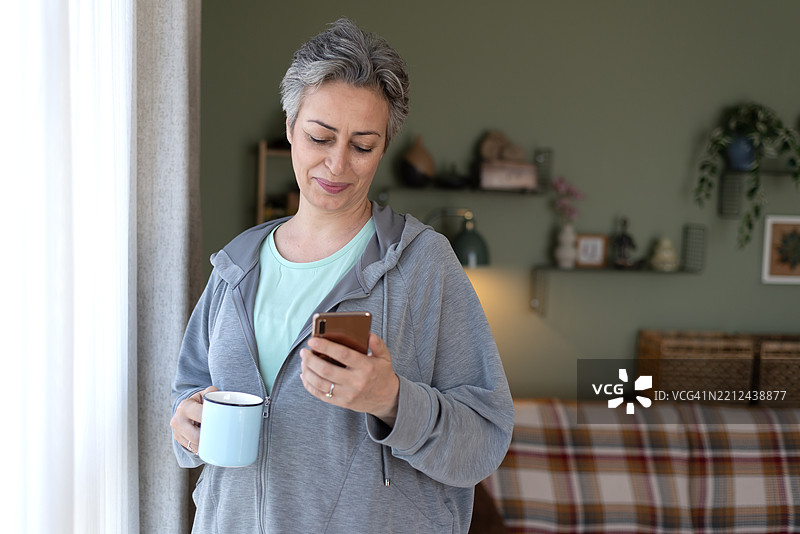 一位成年女性站在窗前，一边喝咖啡一边使用手机。图片素材