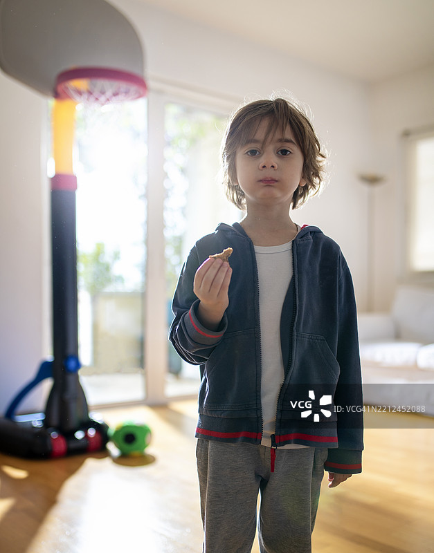 孩子站在明亮的客厅里，手里拿着零食，靠近篮球架。图片素材