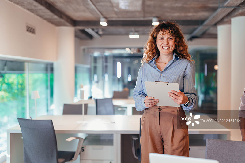 微笑的女商人手持夹板站在现代办公室里图片素材