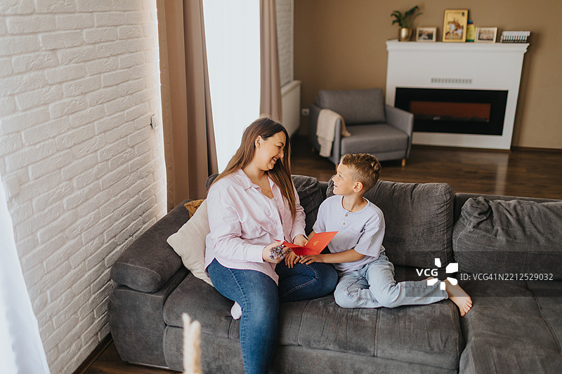 感动的母亲坐在沙发上，拥抱着她的儿子，阅读他为母亲节制作的卡片。图片素材