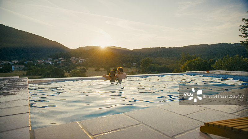 情侣在日落时分的无边泳池中放松，背景是山景图片素材