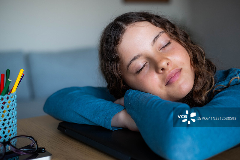 快乐的少女趴在桌子上睡觉，靠在笔记本电脑上，疲惫地休息，戴着眼镜。图片素材