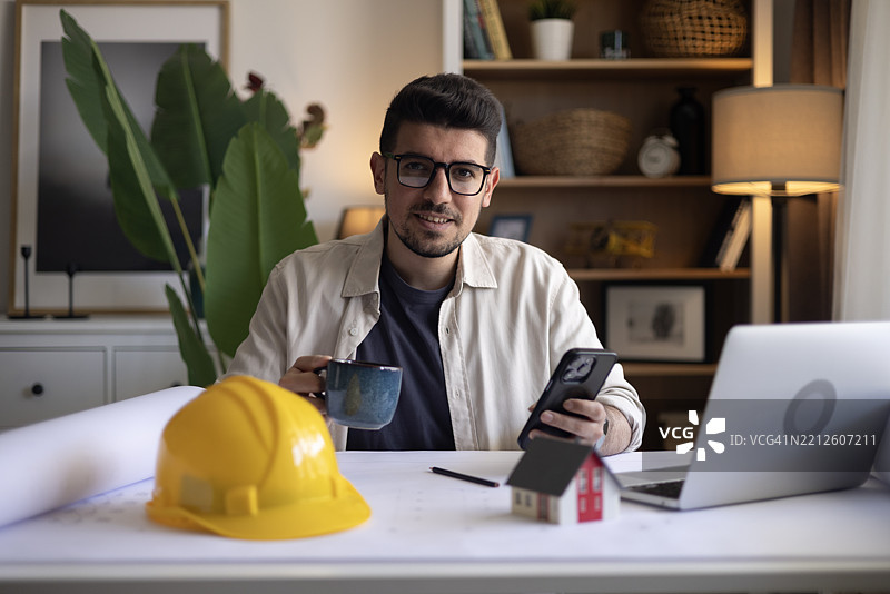 微笑的建筑承包商在家中办公室分析计划。图片素材