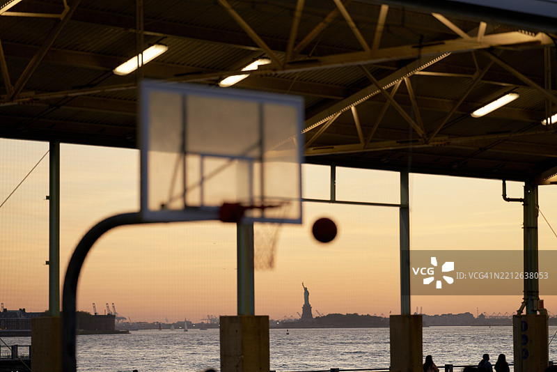 日落时分，篮球架和篮球的剪影映衬着远处的自由女神像。美国纽约市图片素材