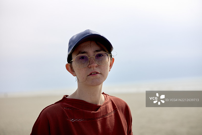 一位戴着帽子和眼镜的女性站在沙滩上，头顶是多云的天空。法国诺曼底图片素材