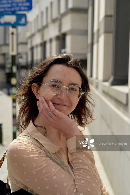 戴眼镜的女性微笑着，手托着下巴，站在城市街道上图片素材