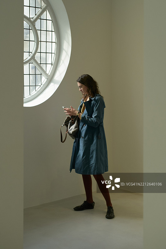 穿着蓝色外套的女性在柔和灯光的房间里，靠近圆形窗户使用智能手机。比利时布鲁塞尔图片素材