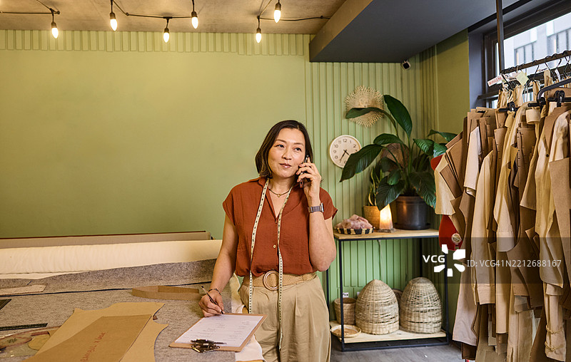 亚洲女性裁缝在店里一边打电话一边做笔记图片素材