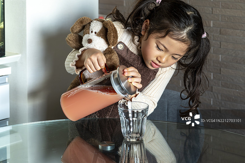 小女孩在家里把果汁倒入杯子，手里抱着一个软玩具。图片素材