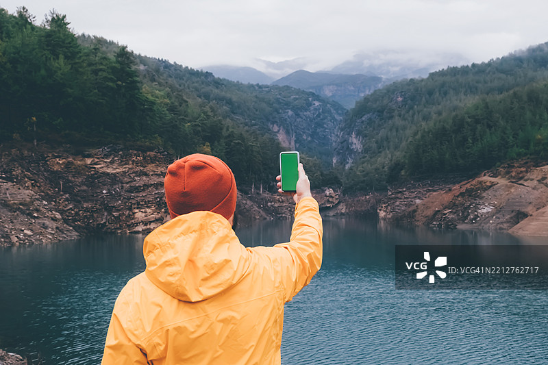 穿着黄色夹克和毛线帽的男子背影在湖边举起手机，周围是松树和远处的山脉。用智能手机探索宁静湖泊的壮丽山景。图片素材