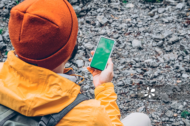 徒步旅行者在岩石环境中利用智能手机技术探索大自然。一位戴着橙色绒帽、穿着徒步夹克的男士背影，手持智能手机，俯视着岩石表面。图片素材