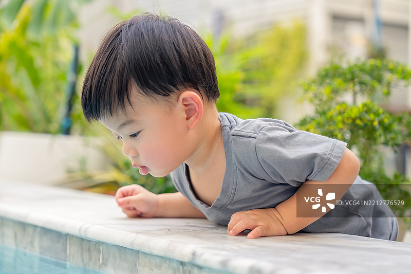 快乐而活泼的亚洲幼儿男孩坐在泳池边，试图探索水面，享受夏季假期的玩耍时光。图片素材