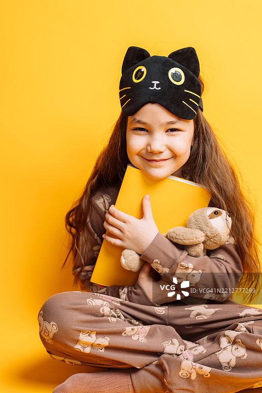 小女孩戴着睡眠面罩，穿着睡衣，抱着她最喜欢的毛绒玩具和书，背景为黄色。图片素材