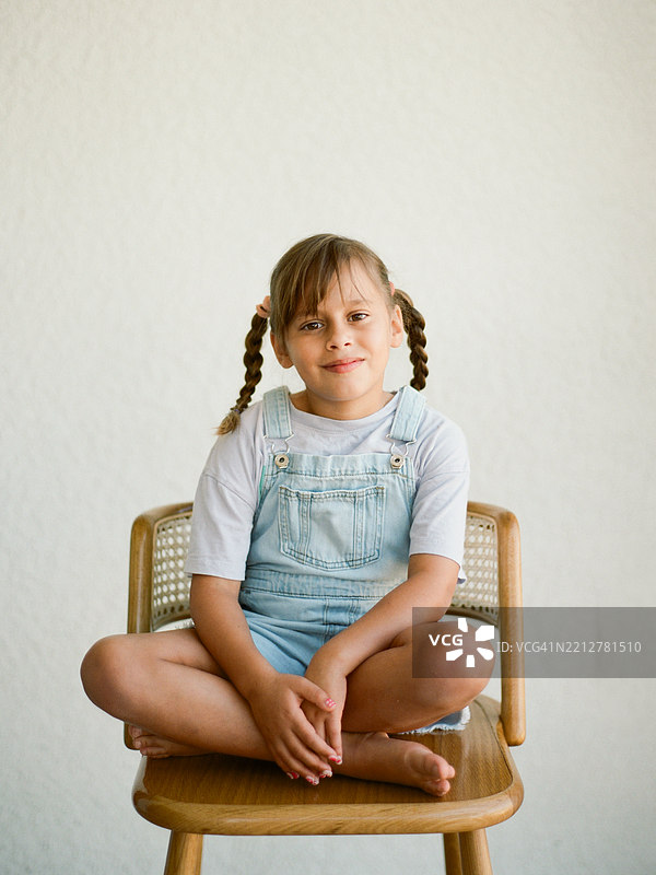 一个6到8岁的小女孩，扎着辫子，穿着工装，站在干净的白色墙壁前。图片素材