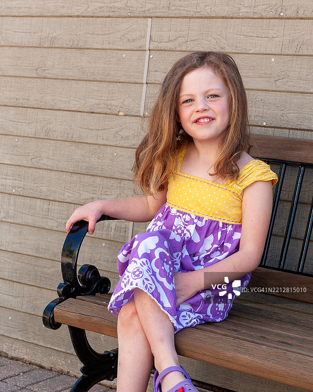 可爱的小女孩坐在长椅上，双腿交叉，微笑着，穿着紫色和黄色的夏装，坐在户外。图片素材