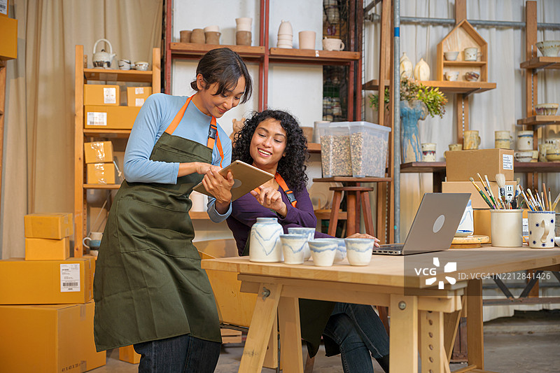 两位女商人在管理，女性正在检查库存并更新库存信息，另一位则在查看客户的在线订单。电子商务业务伙伴在仓库中共同为在线业务工作。图片素材