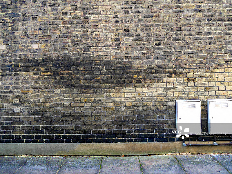风化且肮脏的砖墙上有两个小柜子和燃气管道（经过修饰，无标志或商标）。铺砌的人行道。伦敦，英格兰，联合王国。图片素材