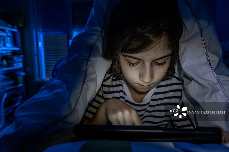 小女孩在晚上躲在毯子下用平板电脑。图片素材