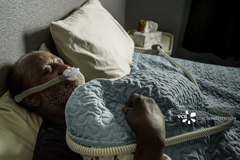 一位七十多岁的非裔美国老人安静地躺在床上，使用CPAP（持续正压呼吸）机来缓解睡眠呼吸暂停。图片素材